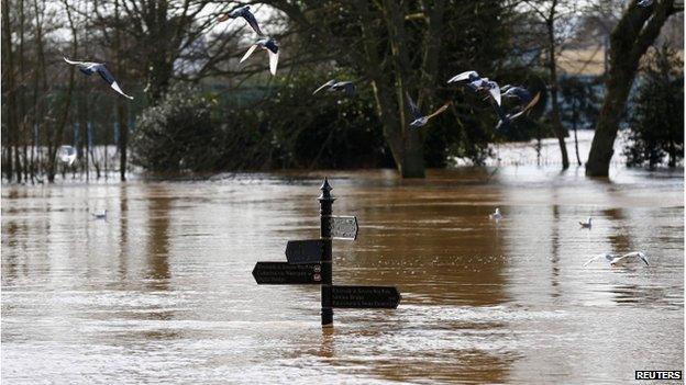 Birds fly past a sign surrounded by flood waters in Worcester