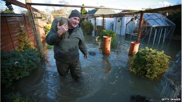 A man carries a sandbag on his shoulder through a flooded garden