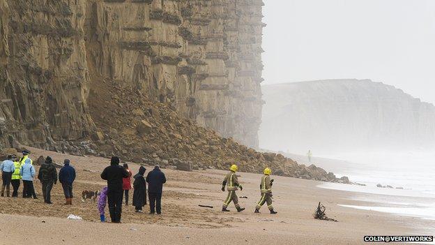 Firefighters and members of the public stand on a beach, looking at a collapsed section of a cliff