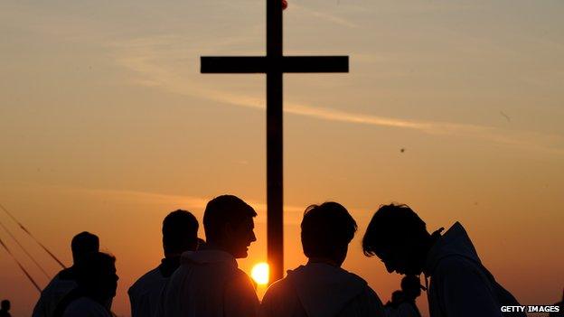 Priests stand by a crucifix with the sun setting behind