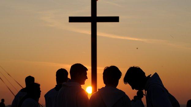 Priests stand by a crucifix with the sun setting behind