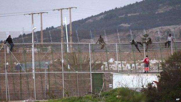 Sub-Saharan migrants climb over a metallic fence that divides Morocco and the Spanish enclave of Melilla, as a Red Cross worker is on-hand to offer humanitarian assistance (17 February 2014)