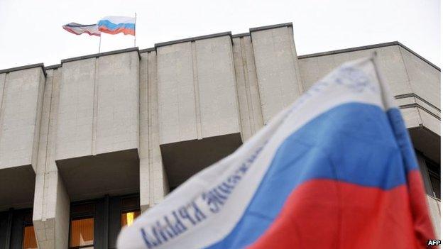 Russian and Crimean flags flutter on top of the Crimean parliament building, with another Russian flag in the foreground