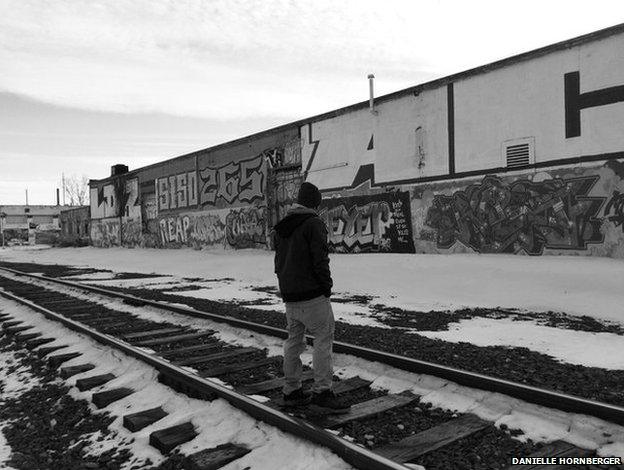 A man stands with his hands in his pockets, looking at a wall covered in graffiti