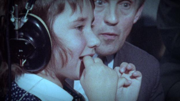 A young girl has headphones on and is watching something out of shot