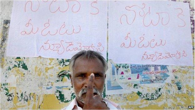 An Indian voter poses with his ink-marked finger in front of posters saying vote for None Of The Above