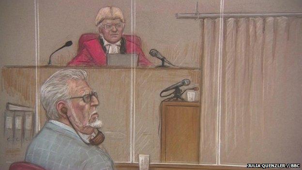 Rolf Harris watches on from the dock as a witness gives evidence behind screen at Southwark Crown Court, watched by the judge, Mr Justice Sweeney