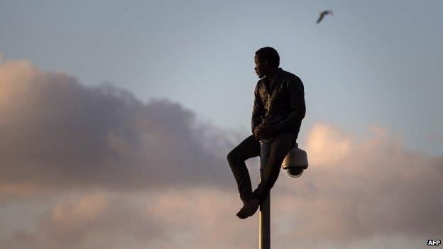 A sub-Saharan migrant sits on top of a pole set in a metallic fence that divides Morocco and the Spanish enclave of Melilla