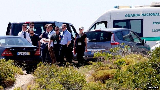 A group of people wearing ID badges standing near parked cars and vans, including a police van