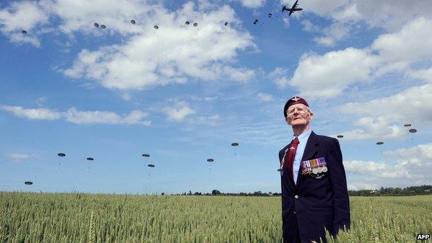 Veteran Frederick Glover posing for a photograph as soldiers parachute in Ranville