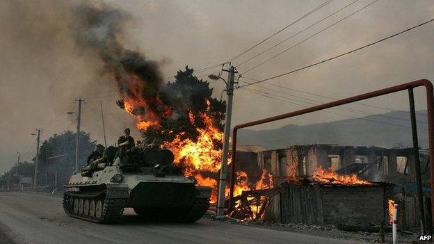 A Russian armoured troop-carrier moves with soldiers (troops) atop next to a house set on fire by South Ossetian militia