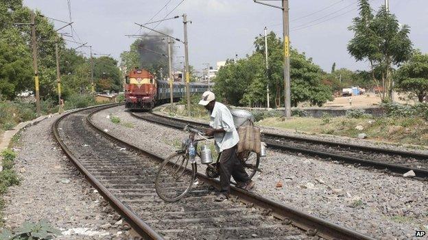 An Indian snacks vendor crosses railway tracks with his bicycle as a train approaches, behind, in Hyderabad, India