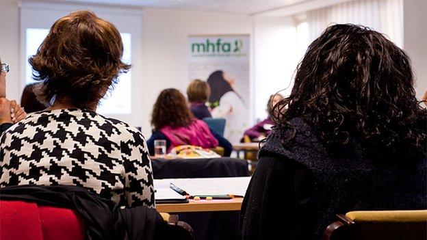 An MHFA training classroom with people sitting at desks listening to a talk