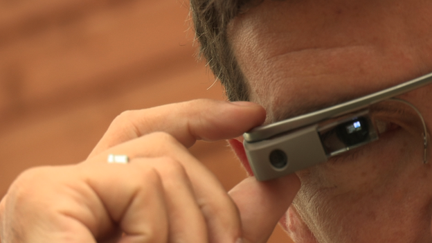 A close up of man wearing the Google Glass. The glass screen goes directly across his eye and his is pressing a button on the top of the glass