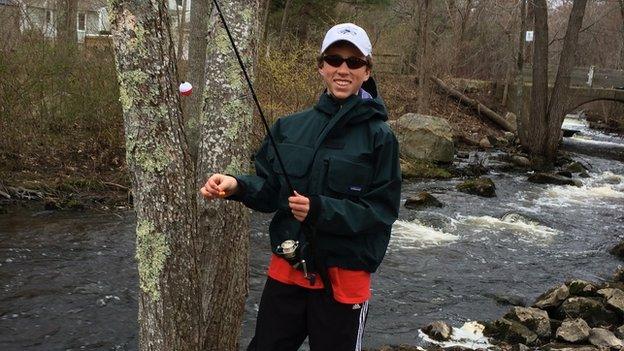 Christopher Herndon, an adolescent boy, is in a river wearing fishing gear and holding a fishing rod.