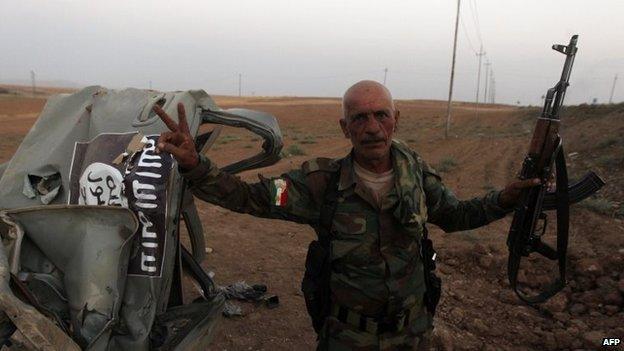 A peshmerga fighter flashes the sign for victory next to the remains of a car, bearing an image of the jihadist flag