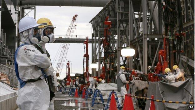 Workers stand by as others handle machinery and pipes which will be used to create a frozen underground wall to surround the crippled reactor buildings at Tokyo Electric Power Co.'s Fukushima Daiichi Nuclear Power Plant in Okuma, Fukushima Prefecture, northeast of Tokyo, Japan, Wednesday, 9 July 2014.