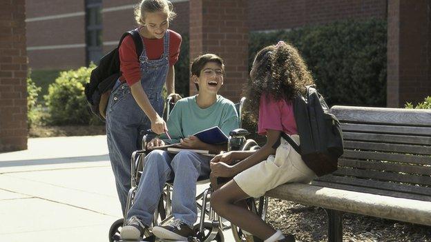 A boy in a wheelchair with two female friends, one of them is sitting on a bench