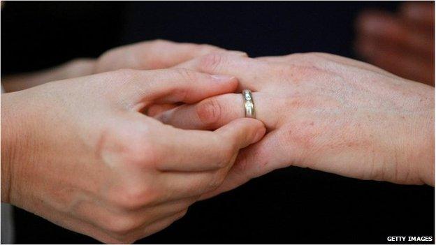 A close-up of hands as one person puts a wedding ring on the finger of another