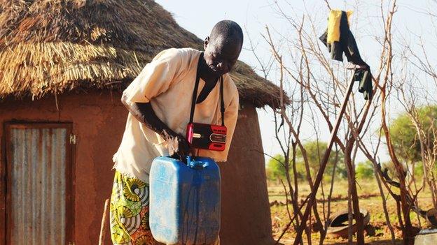Man with portable radio hanging from his neck, carrying a water can in front of a mud hut