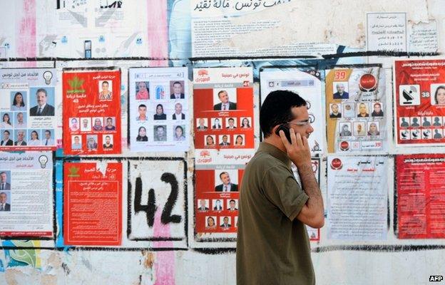 A Tunisian speaks on the phone as he walks past election posters put up on a street ahead of the parliamentary election in the Tunis suburb of Ariana on October 21, 2014.