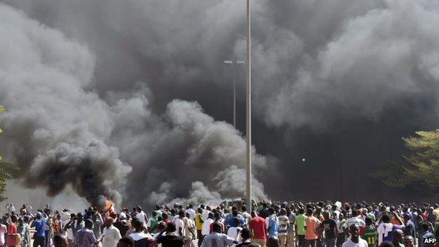 People stand in front of smoke rising from the Burkina Faso's parliament, where demonstrators set cars on fire parked in a courtyard of the Parliament, in Ouagadougou, 30 October 2014