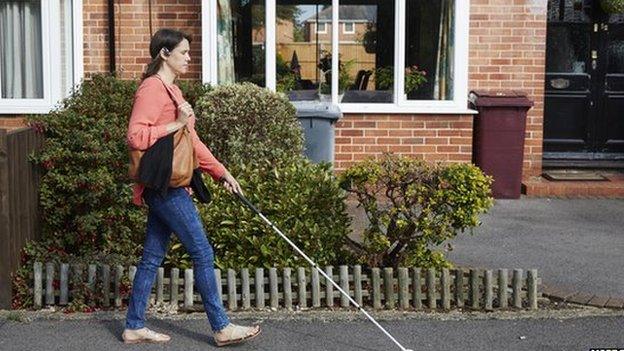 A woman with a white stick and the headset walking down the street