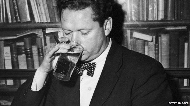 Welsh poet Dylan Thomas (1914 - 1953) drinking a glass of beer and smoking while seated at a desk with stacks of his books of poetry, New York City, c. 1950.