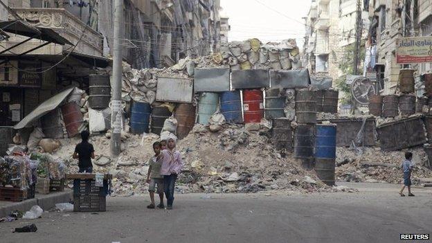 Children walk in front of barrels and sandbags stacked up in a makeshift cover for protection against sniper fire in the East Al-Ansari neighborhood of Aleppo on 7 September 2014