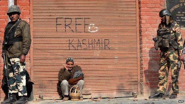 Indian paramilitary soldiers look on as a Kashmiri resident leans against a shuttered storeroom for a smoke in Tral.