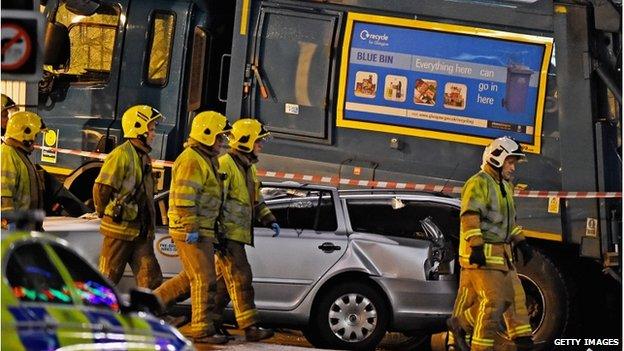 Firefighters walk close to a crashed bin lorry and a damaged car