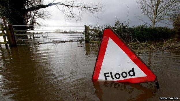 A flood sign is seen in flood waters surrounding farm buildings