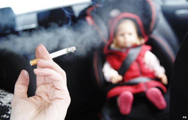 Hand holding cigarette in front of a baby or doll in a car seat