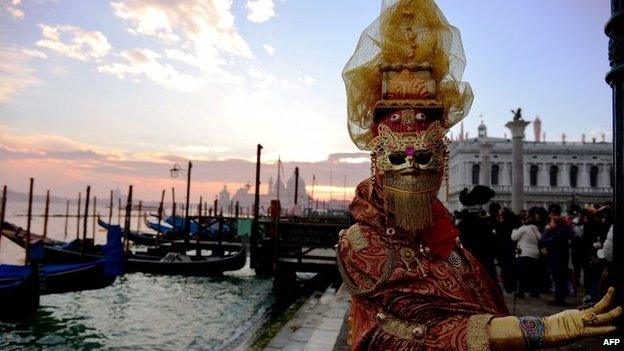 A costumed reveller poses in front of gondolas during the Venice Carnival on February 8, 2015 in Venice