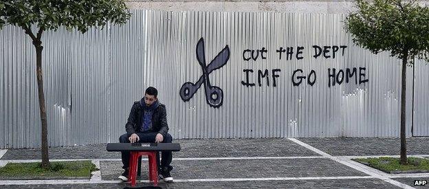 A man plays music in front of a sign saying IMF Go Home, Athens, 24 Feb 2015