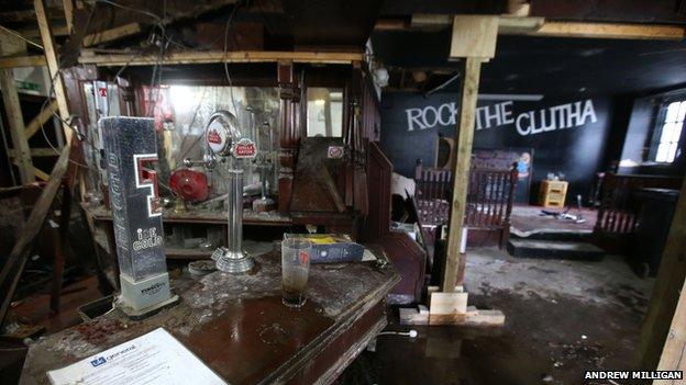 A pint glass and change on the bar; a Tennants tap. View of stage 'Rock the Clutha' backdrop