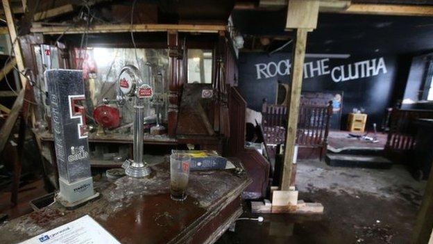 A pint glass and change on the bar; a Tennants tap. View of stage 'Rock the Clutha' backdrop