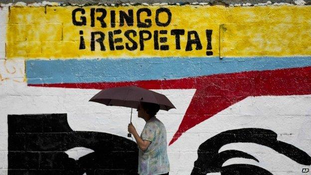 A woman using an umbrella during a drizzle walks by a mural representing the eyes of Venezuela's late President Hugo Chavez that reads in Spanish "Gringo, respect!" in Caracas, Venezuela, Monday, March 2, 2015.