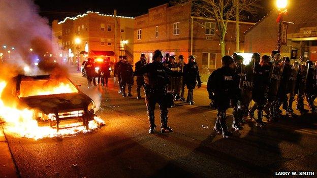 Row of police in riot gear beside a burning car