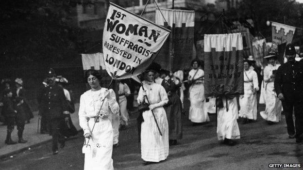 A protest march by women suffragettes in London with police in attendance. The banner held by the leading women reads "1st Woman Suffragist Arrested in London"