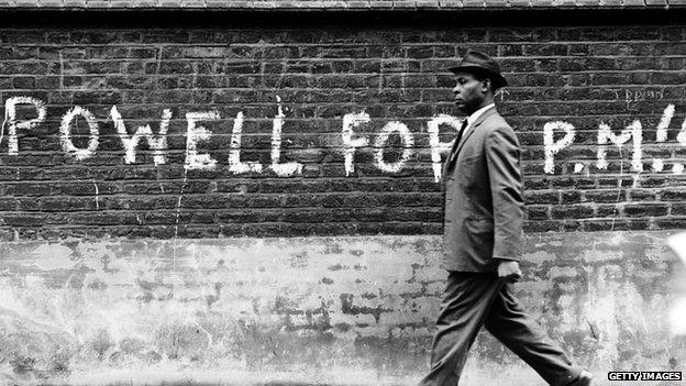 A black man walking past graffiti stating "Powell For PM" in 1968