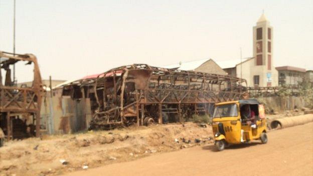 A motorised tricycle passes the wreckage of burnt out busses in Kano, Nigeria