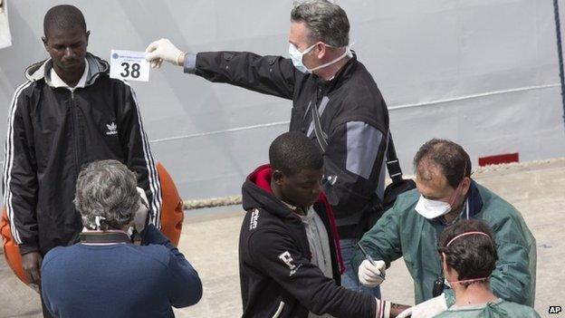 An Italian forensic police officer holds a numbered tag as another one takes a photo of a man who disembarked from the Italian Coast Guard ship " Fiorillo " at the Catania harbor, Sicily, southern Italy, Friday, April 24, 2015.