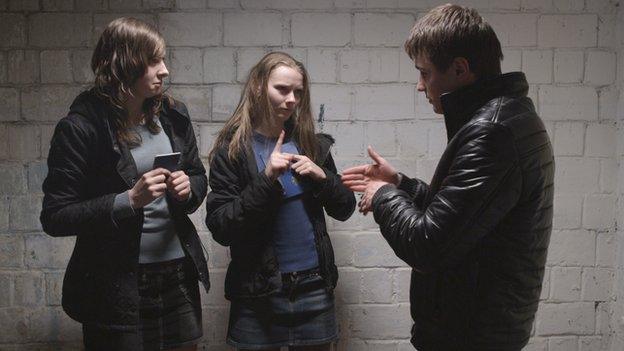 Two women and a man signing to each other against a white brick wall