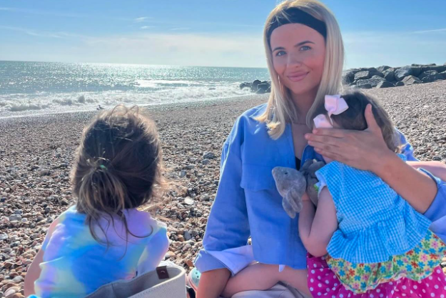 A woman is sat on a pebbled beach, smiling at the camera. She has a child next to her and is holding a toddler on her knee. 