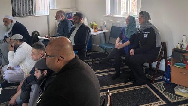 A small group of men sit in a room with white walls.  Some of the men are wearing skull caps. Two women sit at the back - bother are wearing blue scarves over their heads (Danielle Stone and Sophia Parveen)