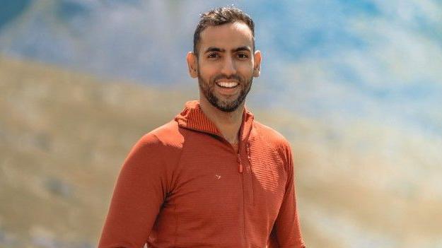 Sameed Asghar is standing in front of a hillside which is blurred in the background. He has short dark hair and dark facial hair. He is wearing a orange top