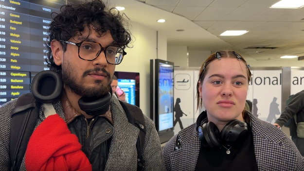 A man and woman stood in an airport terminal. They are both wearing grey jackets and a looking off to the right of the camera, with plain expressions on their faces