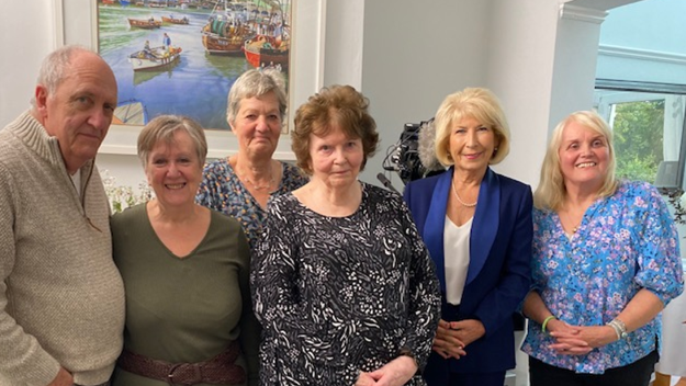 Six grandparents, including Jane, standing in a home together in a group, all looking to camera with a picture of a river and boats on the wall behind. Jennie Bond, the former BBC journalist and television presenter, is amongst them, wearing a blue trouser suit and string of pearls