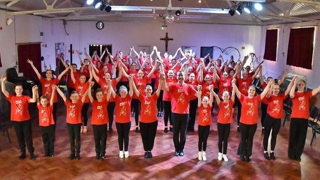 A church hall full of dancers of different ages in red T-shirts with a logo and black trousers.